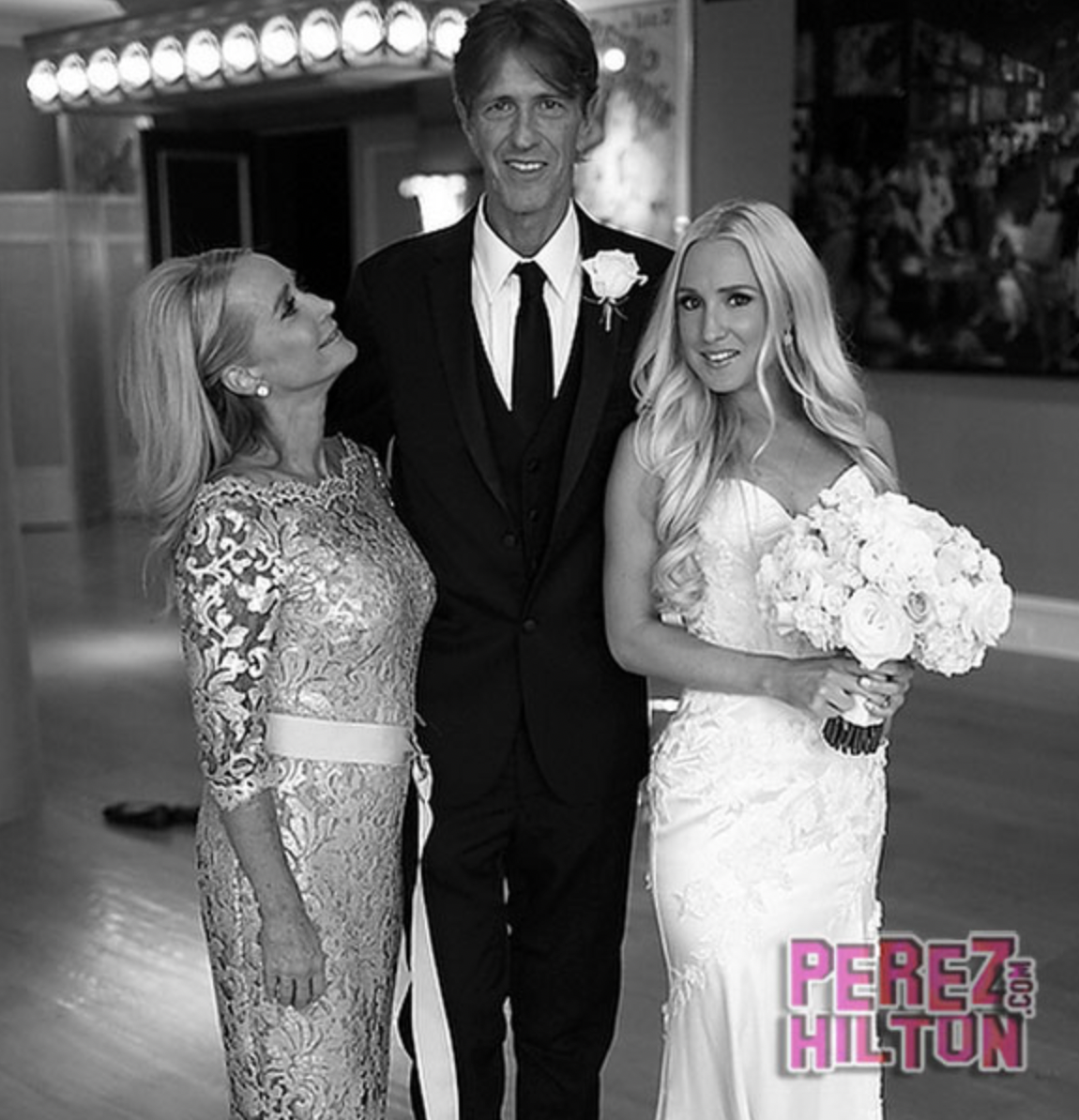 A wedding scene with a bride in a white dress holding a bouquet of roses, a groom in a black suit with a boutonniere, and a woman in a patterned dress looking at the bride, all smiling at an indoor event.