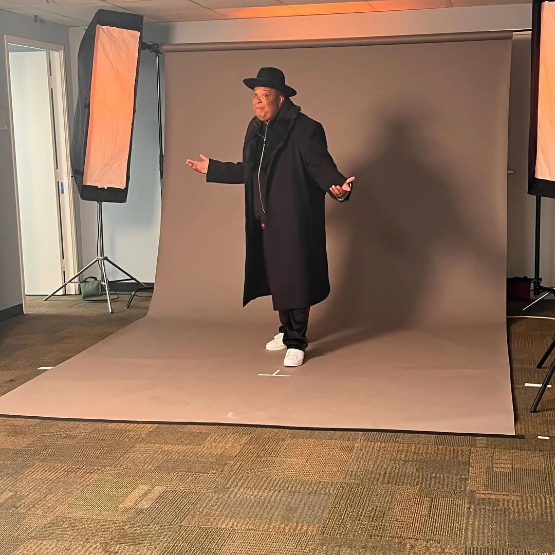 Person standing in a photography studio with a gray backdrop, wearing a black coat, black hat, and white sneakers, gesturing with hands open.