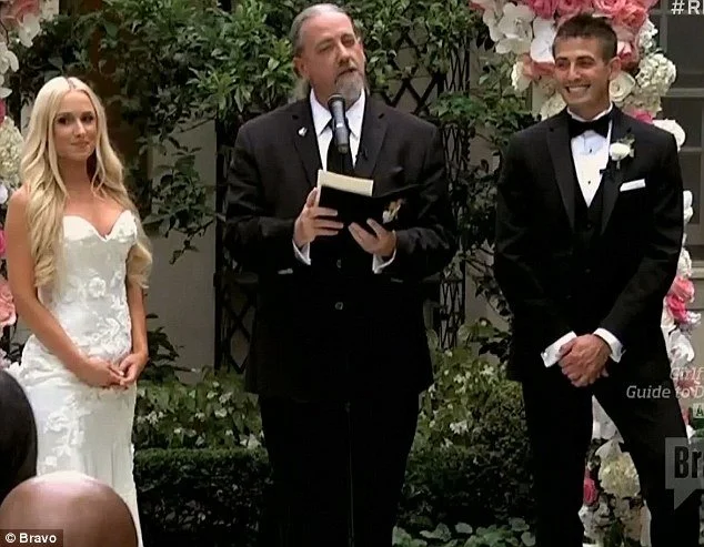 A wedding ceremony with a bride in a white gown, a groom in a black tuxedo, and an officiant in a black suit reading from a book. There are pink and white flowers in the background and greenery.