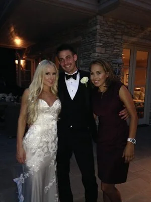 Three people, a woman in a white wedding dress, a man in a black tuxedo, and a woman in a dark dress, standing together at a nighttime indoor event.