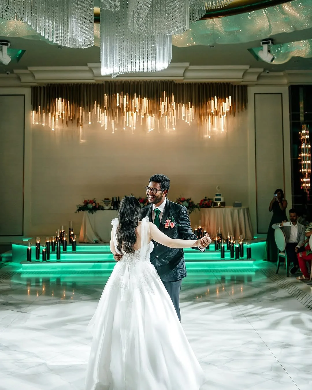 A bride and groom dancing at their wedding reception in a decorated ballroom with a chandelier and green lighting.