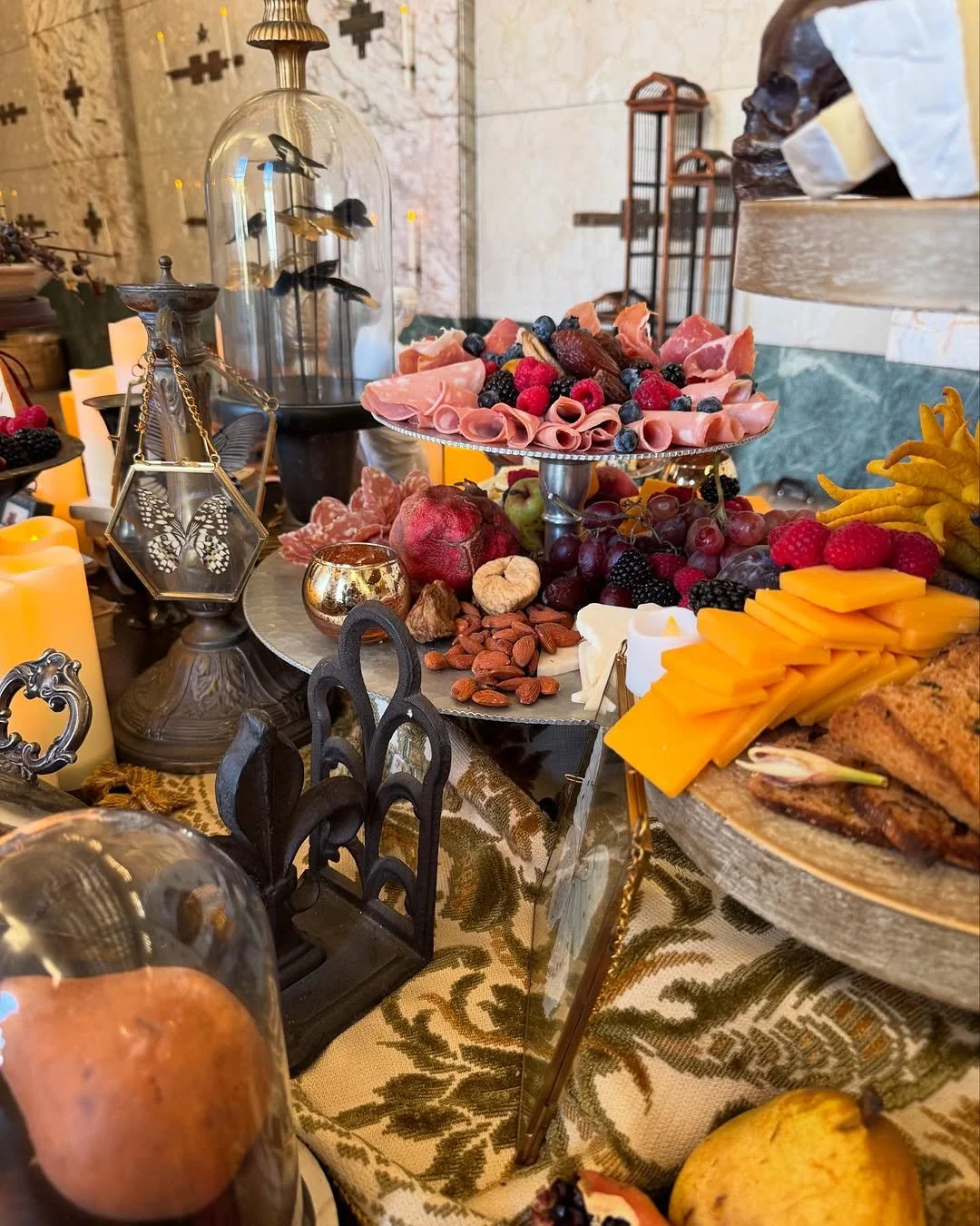 A table set with a variety of cheeses, meats, grapes, berries, and nuts, with decorative candles and a vintage-style display, including a glass dome with flying fish and butterfly ornaments.