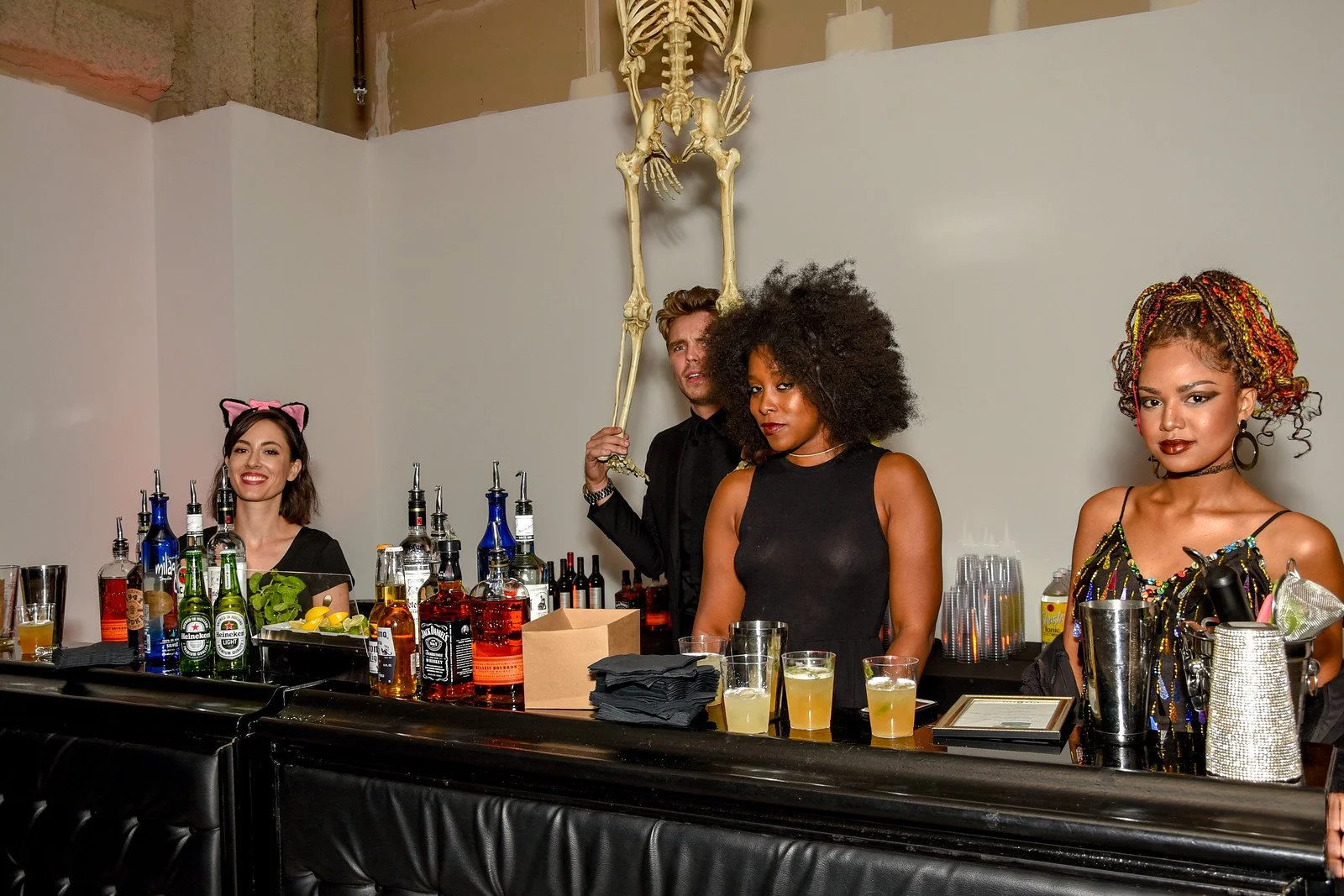 A bar scene with four people: a woman with cat ears headband smiling, a man holding a skeleton model behind his head, a woman with natural curly hair in a black dress looking assertive, and a woman with colorful braids in a sparkly top looking confid