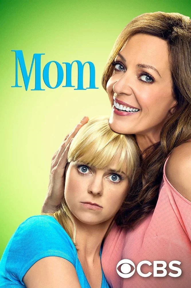 Cover image from the TV show 'Mom' showing two women, one with light brown hair and the other with blonde hair, smiling and hugging against a light green background. The word 'Mom' is written in blue text in the top left corner, and the CBS logo is i