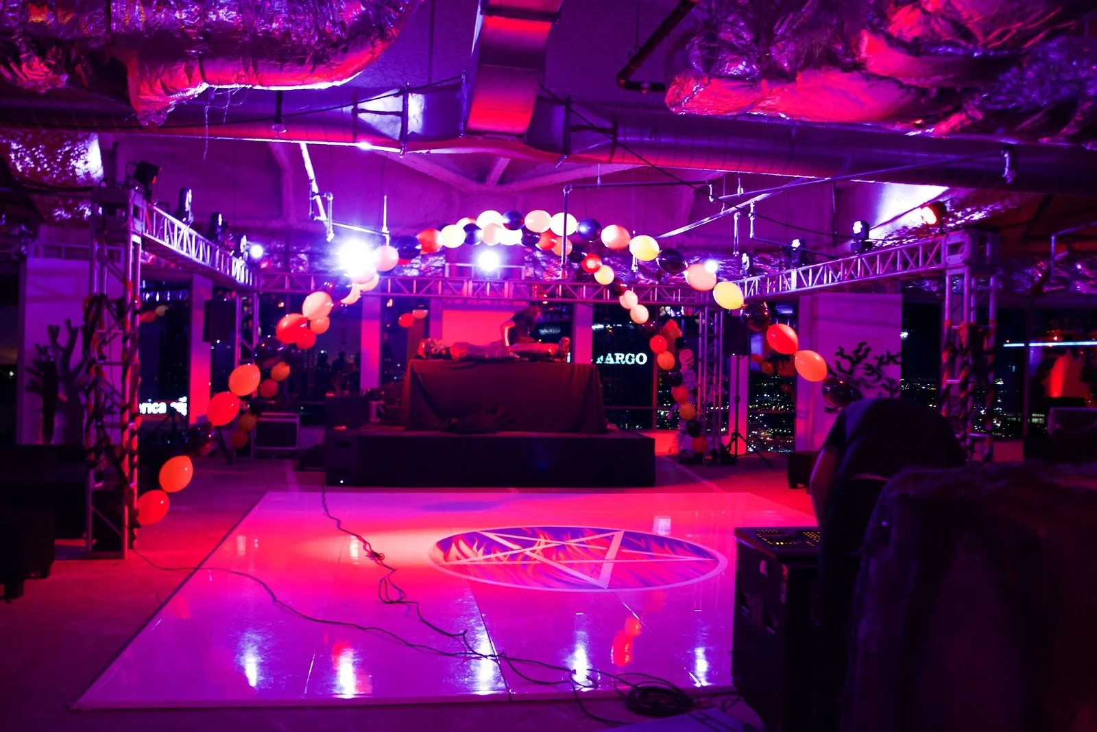 Indoor event space with stage, balloons, and ambient lighting, decorated for a celebration or party, with a city view at night through large windows.