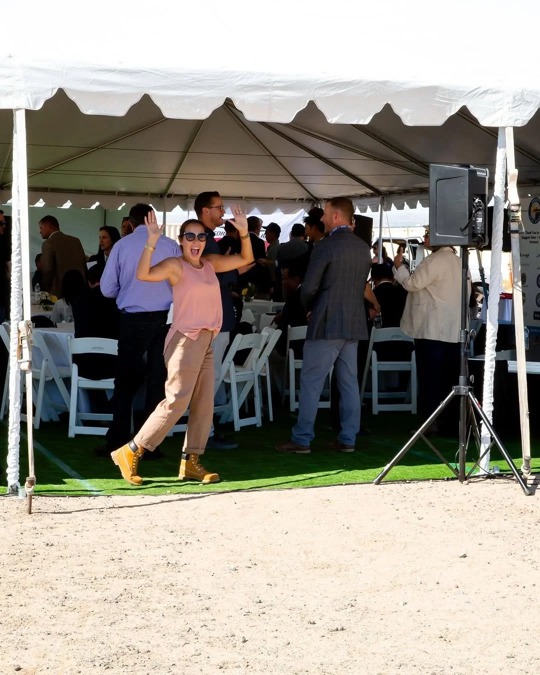 A woman dancing and smiling at an outdoor event under a white tent, with people socializing and sitting at tables behind her.