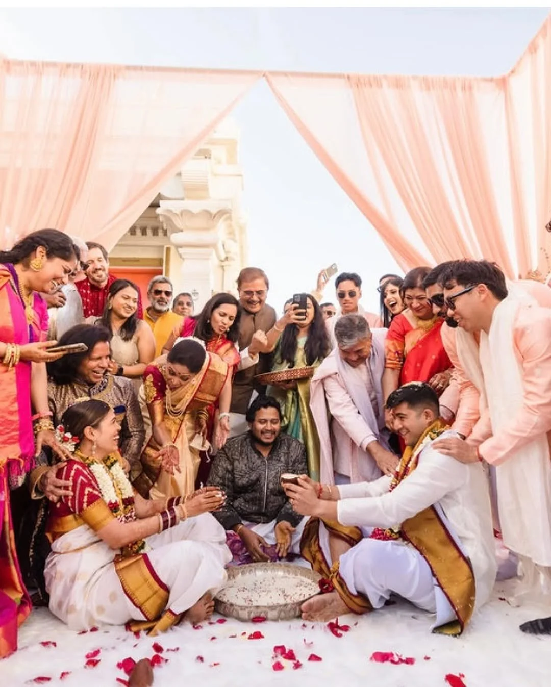 Indian wedding ceremony with bride and groom performing ritual surrounded by family and friends under pink drapes