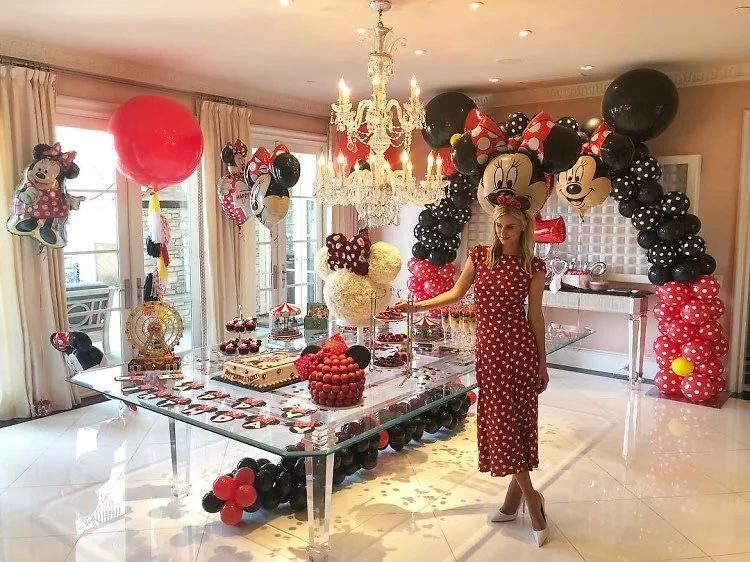Indoor birthday party with Minnie Mouse theme, featuring balloon arch and decorations, a glass table with desserts, and a woman in a red polka dot dress.
