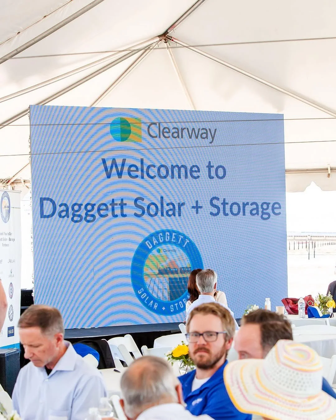 Large screen displays a sign that reads "Welcome to Daggett Solar + Storage" with the Clearway logo above, inside a white tent with tables and people attending.