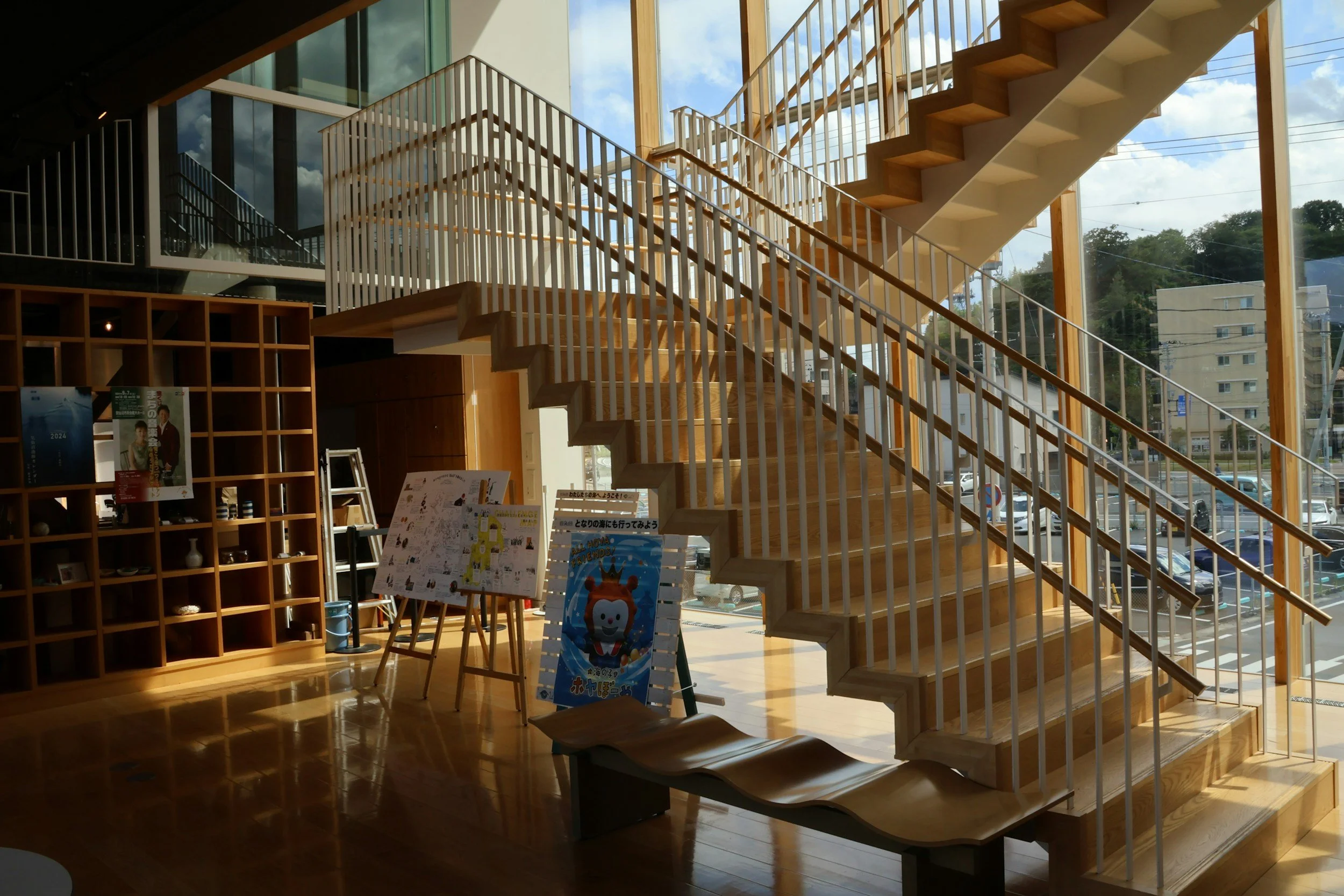 Interior of a modern building with a wooden staircase, large windows, and sunlight streaming in, showing an outdoor parking lot and buildings outside.
