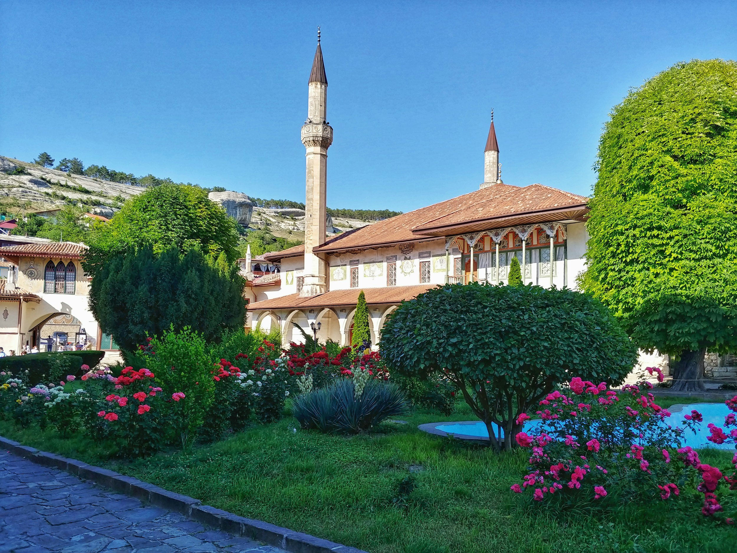 Traditional mosque with two minarets surrounded by a garden of colorful flowers, green bushes, and trees, with a mountain background and a clear blue sky.