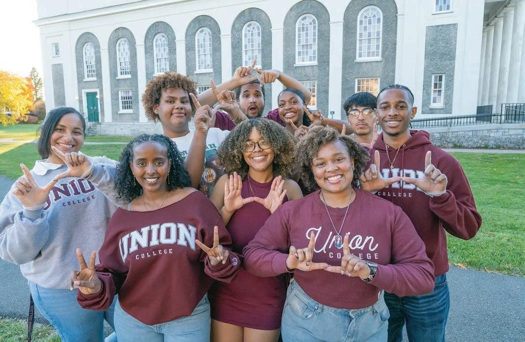 Group of diverse college students posing outdoors in front of a large building, wearing Union College apparel, making various hand gestures and smiling.
