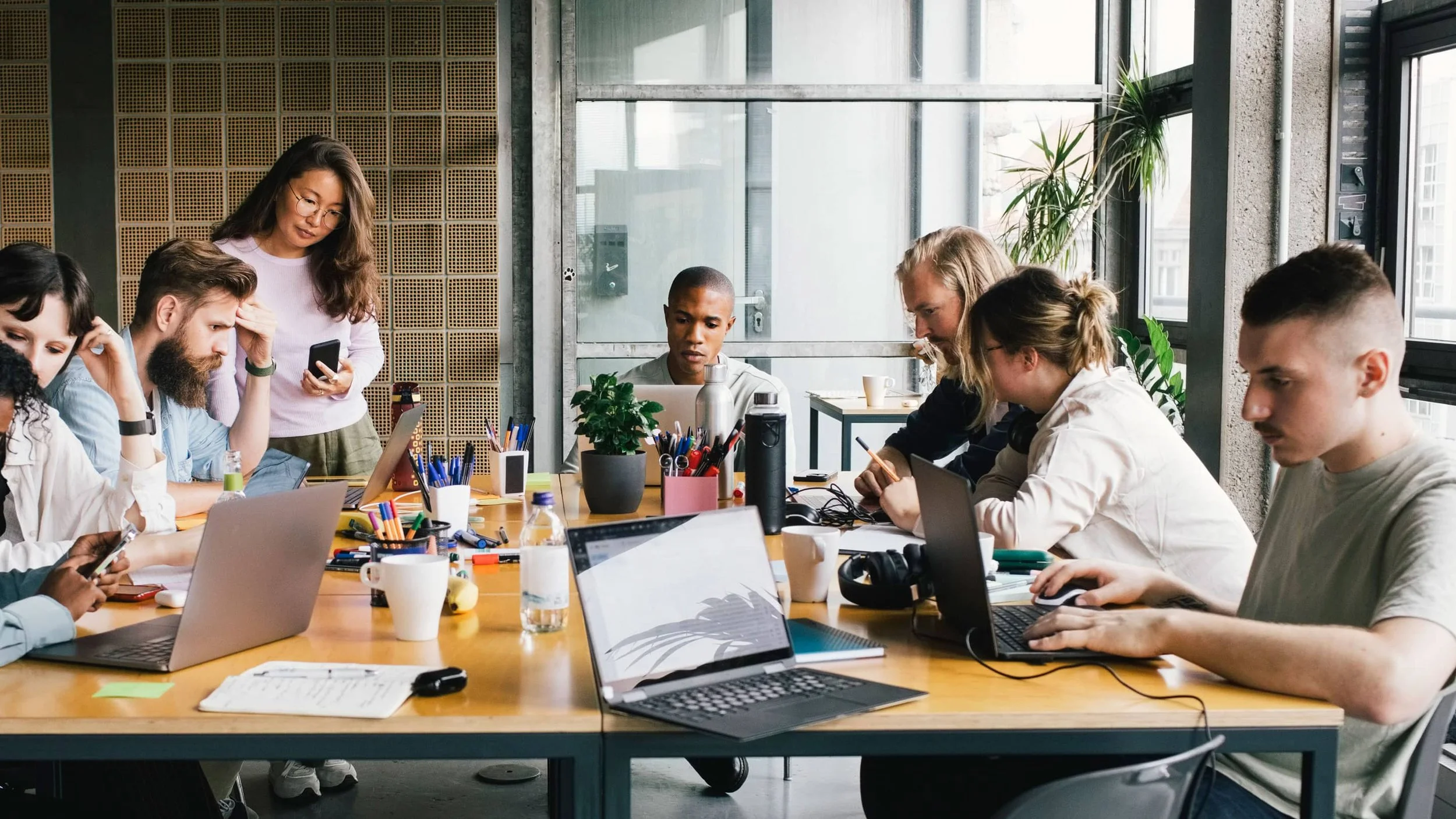 Group of people working on laptops and phones around a large wooden table in a modern office with large windows and potted plants.