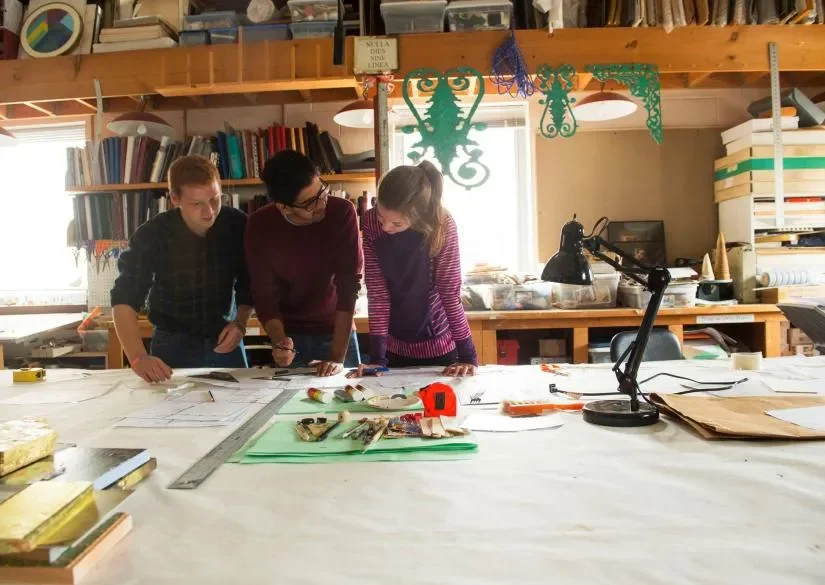 Three people are working on a large table in an art studio, looking at sketches or plans. The table has drawing tools, measuring tape, and papers. The background shows shelves with books and art supplies, and decorative cutouts hang from above.