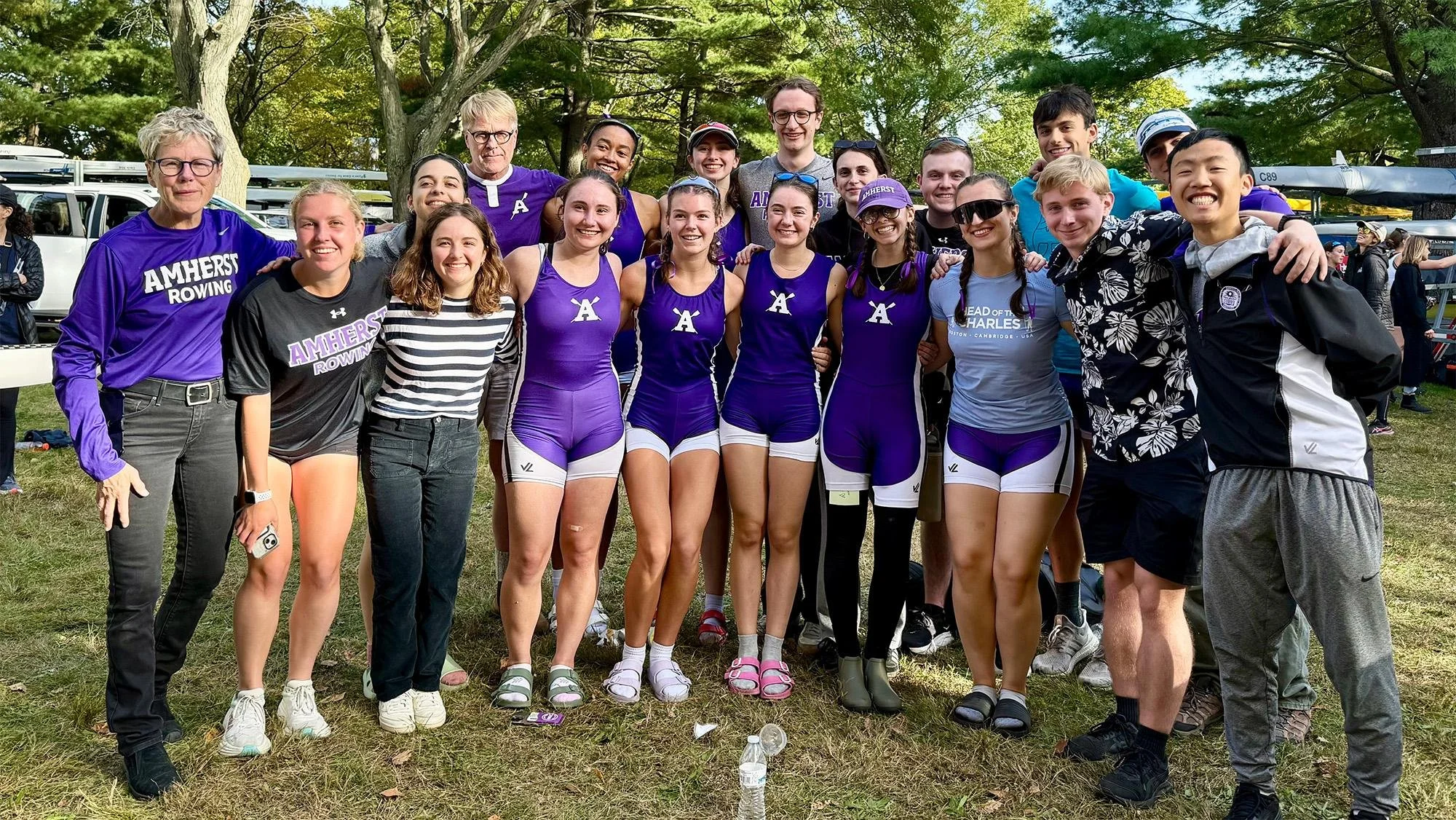 Group of young female athletes and coaches gathered outdoors on grass, smiling for a photo after a rowing event, wearing athletic gear and purple uniforms.