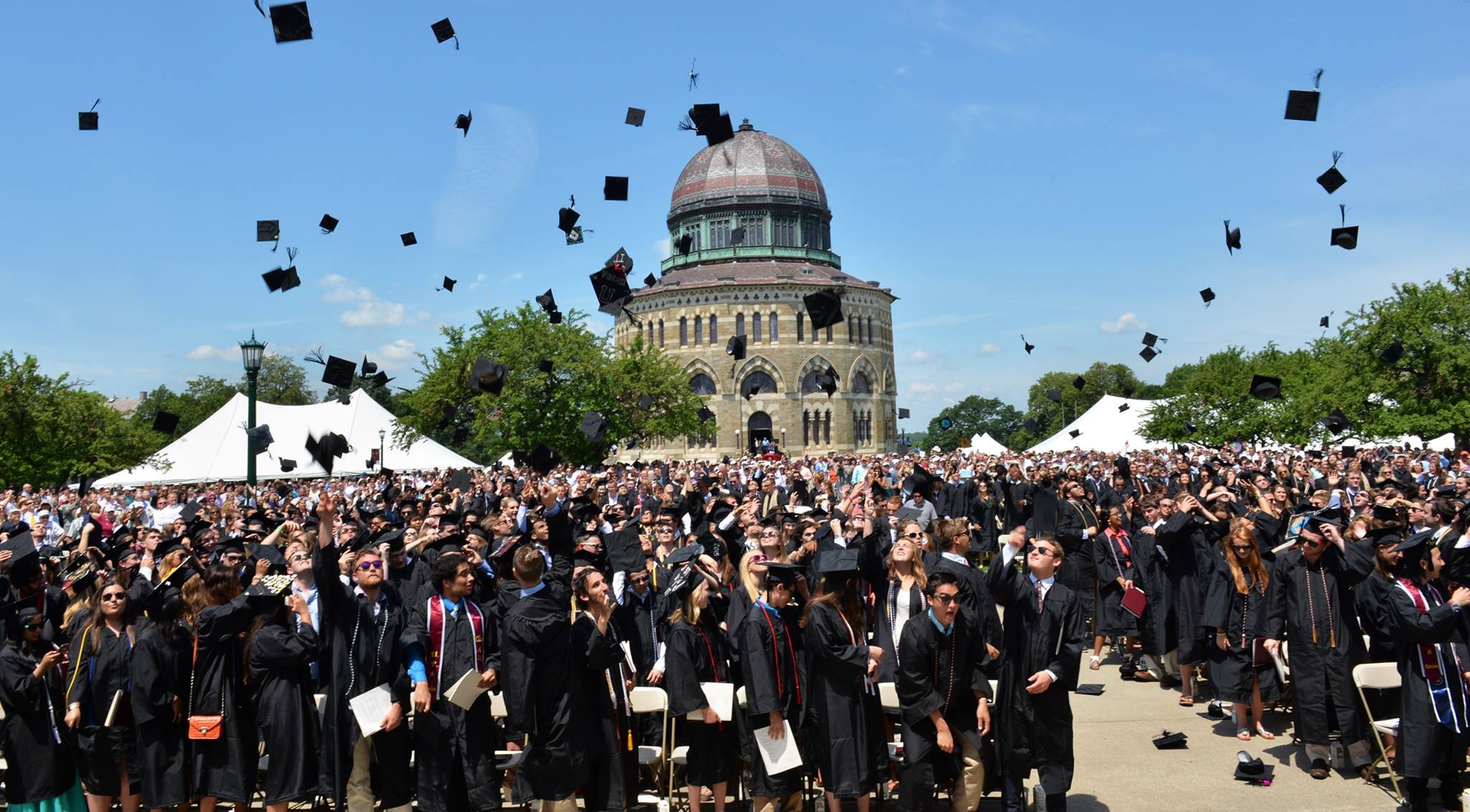 Graduates toss graduation caps into the air during a ceremony on a sunny day in front of a historic building with a domed roof.