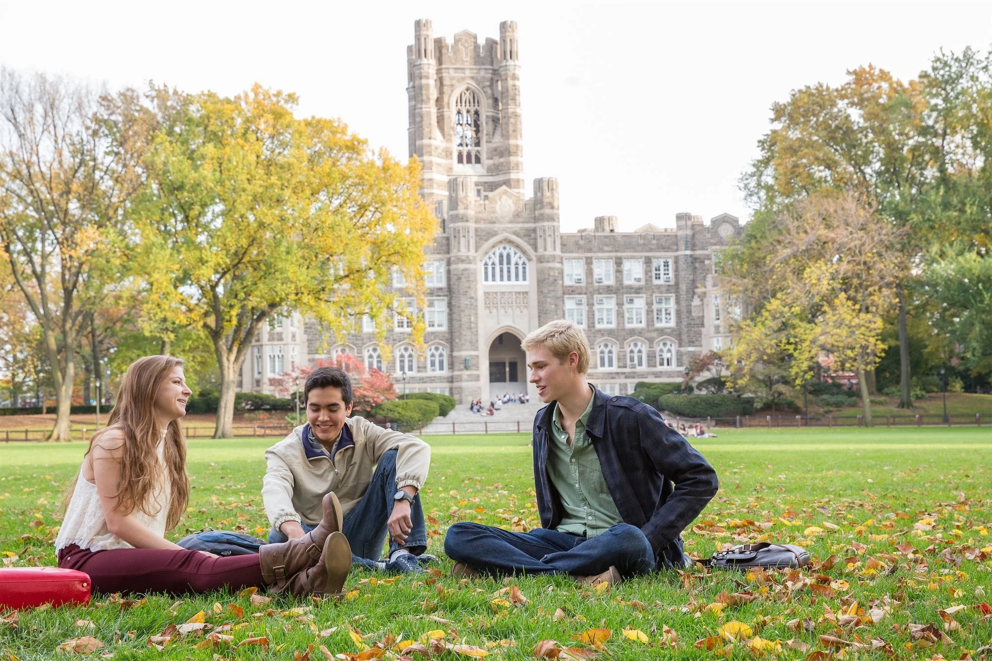 Three college students sitting on autumn leaves on a campus lawn in front of an old university building, laughing and chatting.