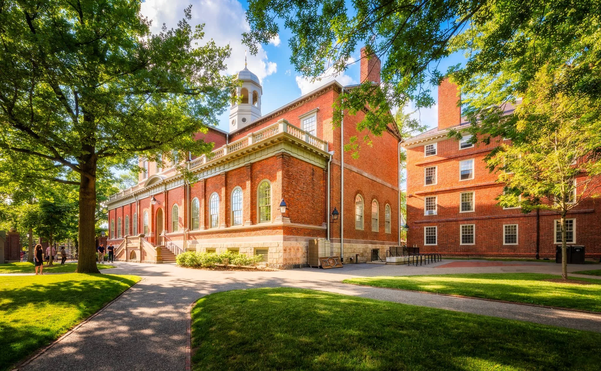 A historic brick building with arched windows, surrounded by lush green trees and a manicured lawn, with a few people walking nearby on a sunny day.