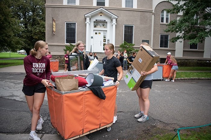 Group of young women moving boxes and items into a large orange cart in front of a residential building.