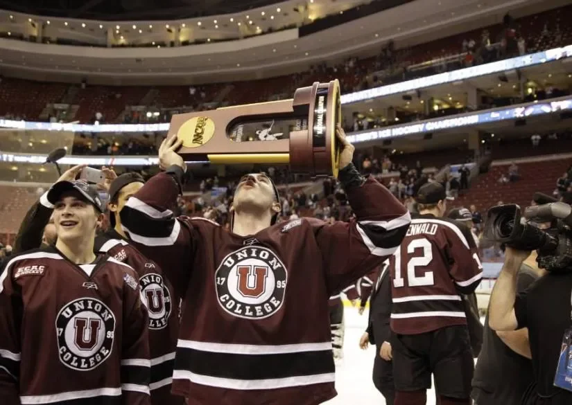 Ice hockey players celebrating victory with one player holding a large championship trophy overhead in an arena.