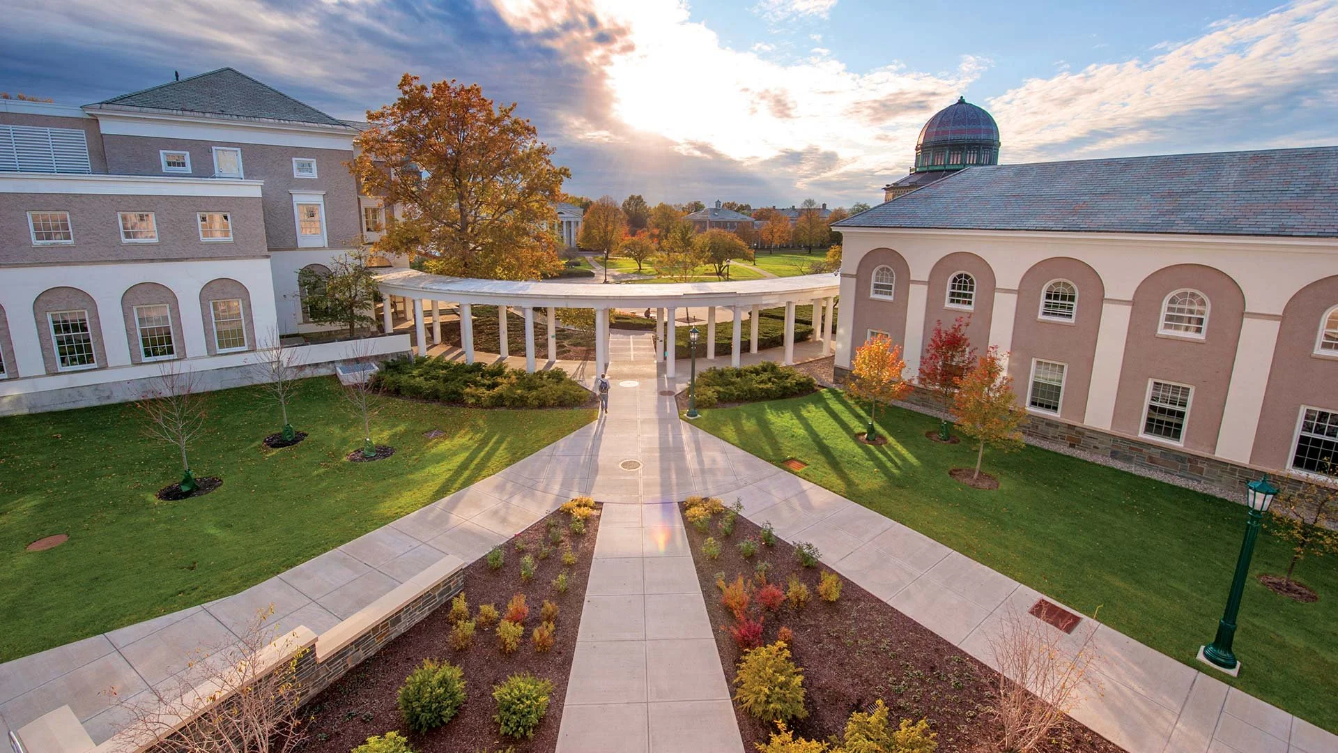 An aerial view of a campus or institutional area with pathways, trees, lawns, classical-style buildings, and a dome structure in the background during sunset.