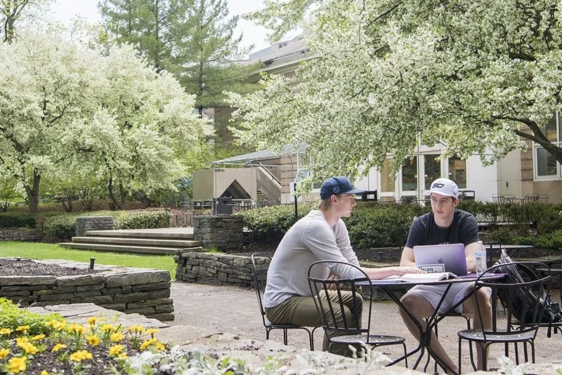 Two young men seated at a black metal table outdoors, engaged in conversation with a laptop and water bottle on the table, surrounded by trees and a brick building in the background.