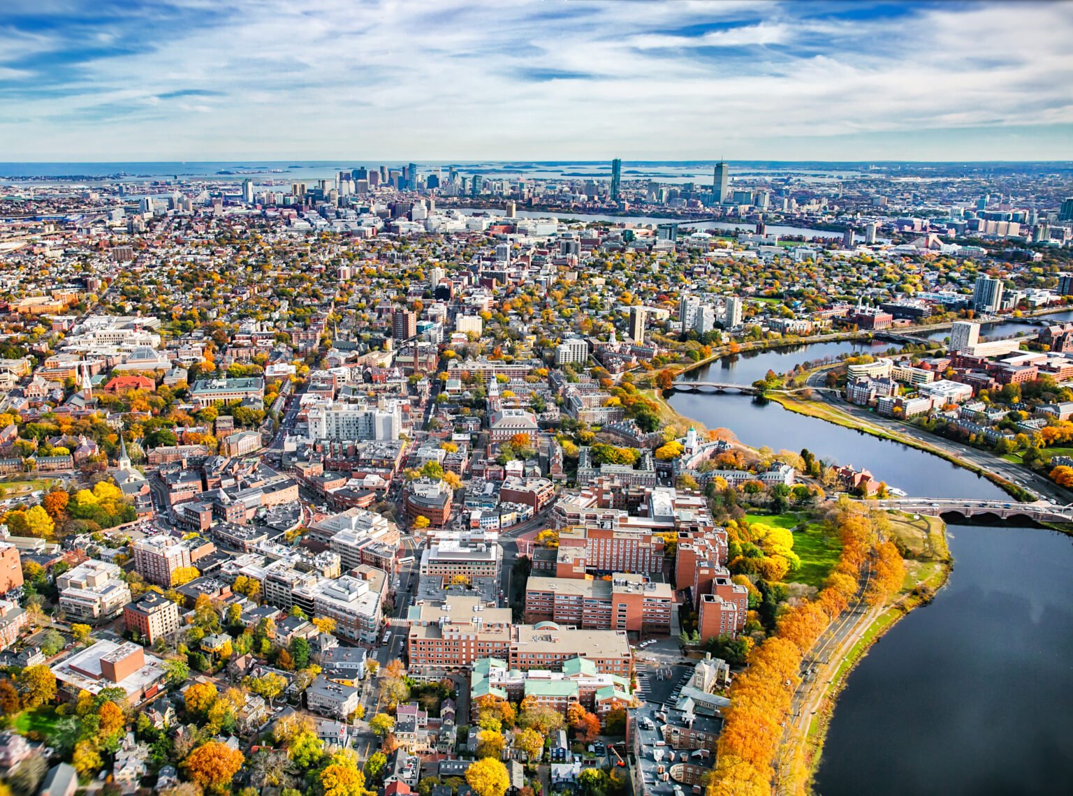 An aerial view of a city with a river winding through it, numerous buildings, and trees with fall foliage.