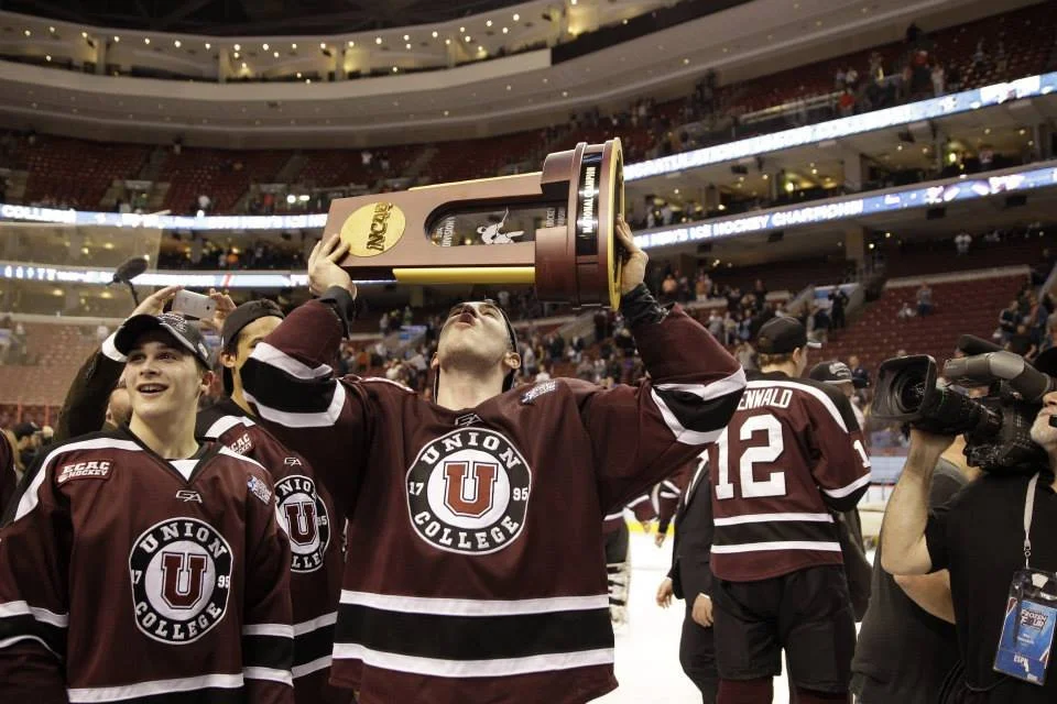 Hockey players in maroon uniforms celebrating a victory, one player is kissing a large championship trophy in a stadium.