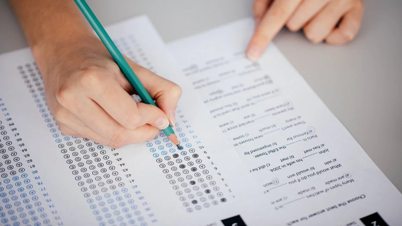 Person filling out a multiple-choice answer sheet with a green pencil, pointing to the 12th question.