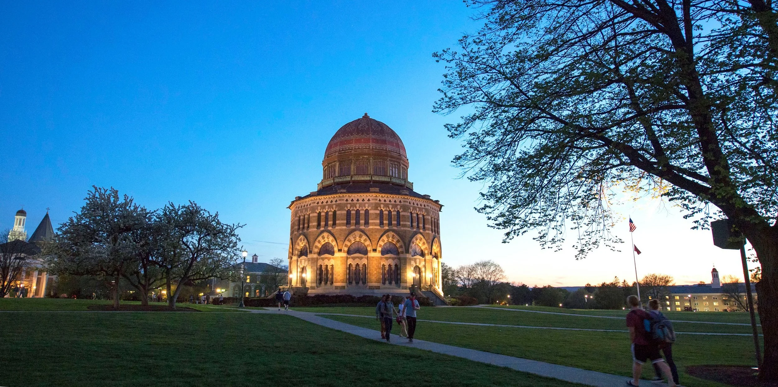 The Texas State Capitol illuminated at dusk with people walking on the sidewalk and trees in the foreground.