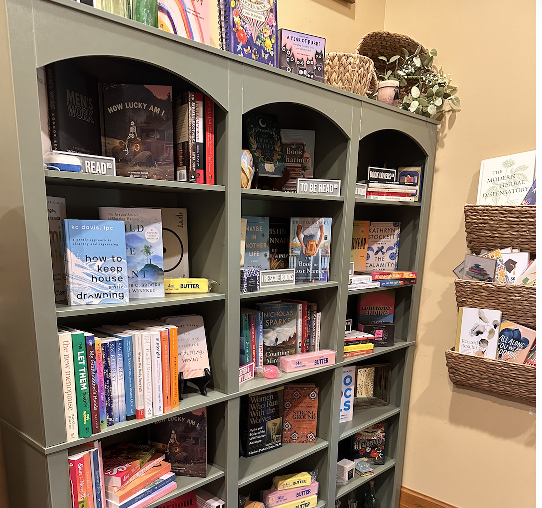 A sage green bookshelf filled with books, some labeled 'To Be Read' and 'Rescue Books.' On top of the bookshelf are decorative baskets, a potted plant, and colorful books. To the right, wall-mounted baskets hold more books and workbooks