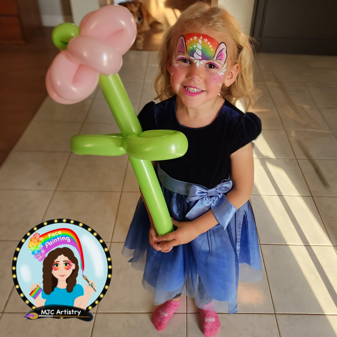 A young girl with face paint of a rainbow unicorn on her forehead, smiling and holding a balloon sculpture of a flower. She is wearing a dark blue dress with a bow, pink socks. Taken at a birthday party in Vancouver BC.