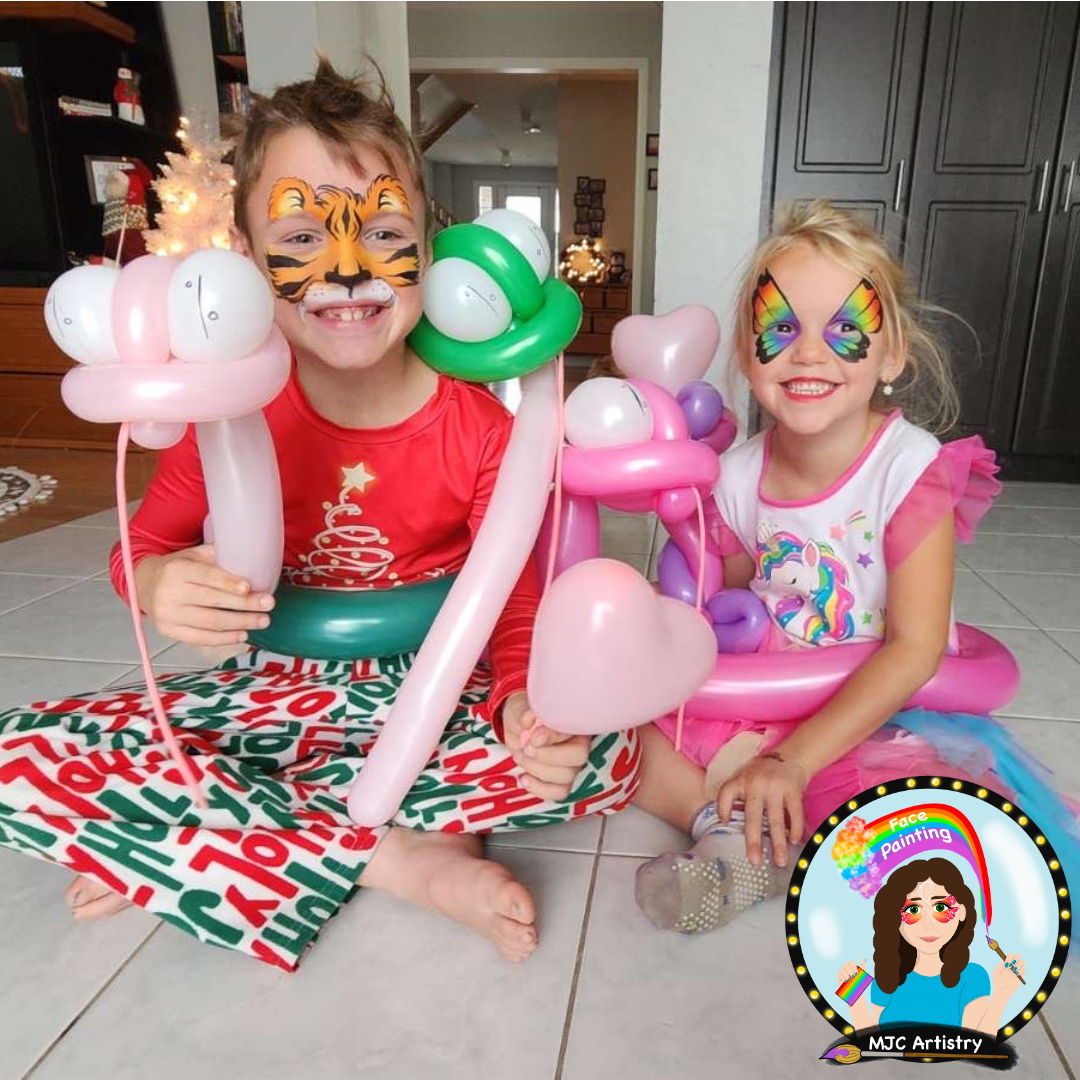 Two young girls with face paint and balloon art, sitting on a tiled floor inside a home. One girl has tiger face paint and a red shirt with holiday design, holding balloons shaped into a tree and other objects. The other girl has rainbow butterfly fa