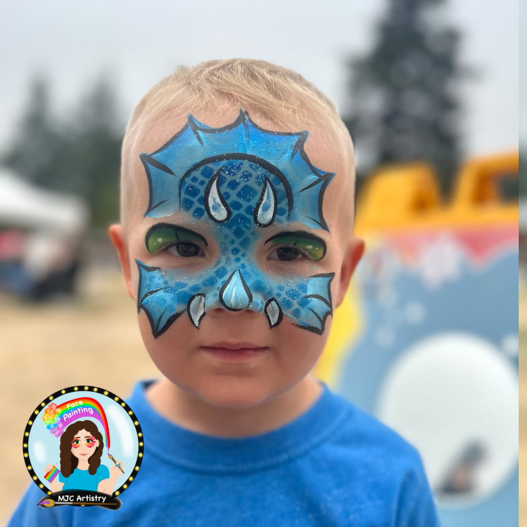 A young child with face paint resembling a blue dragon with green eyes and white scratch marks, standing outdoors with blurred background.