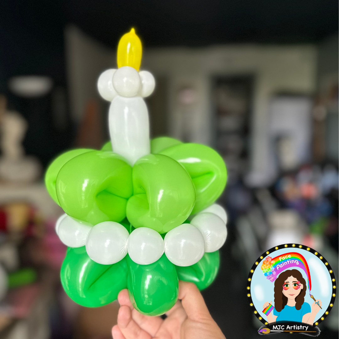 Balloon sculpture resembling a green shamrock with a white candle and yellow flame, held by a hand, against a blurred indoor background with a face painting logo in the corner.