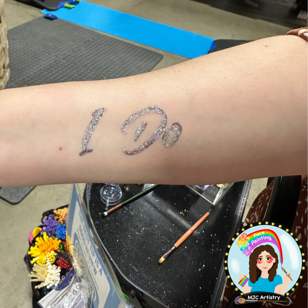 A person has a temporary glitter tattoo on their forearm that reads "I Do". There are makeup brushes and colorful accessories nearby, suggesting face painting or body art activity in progress.
