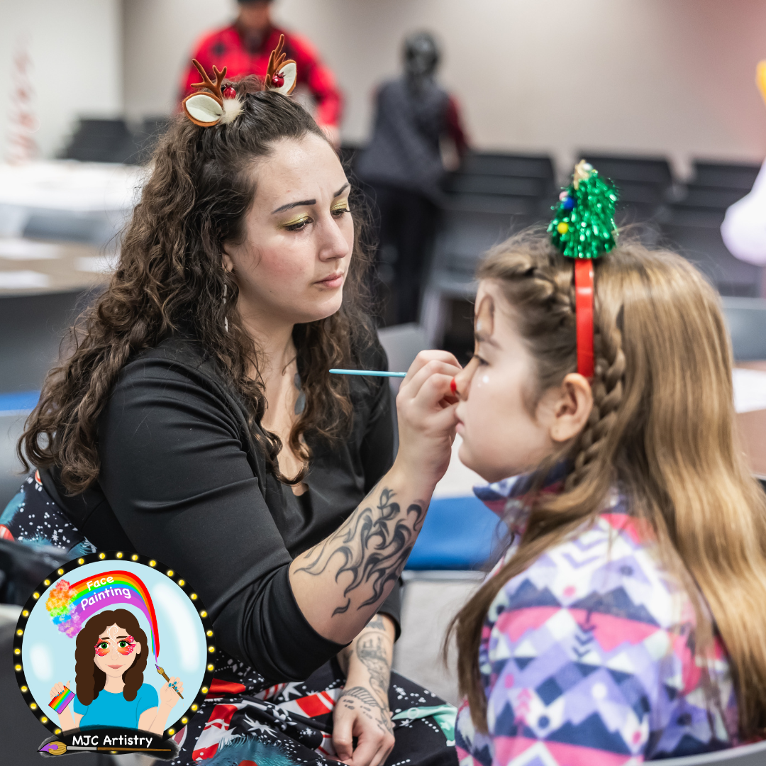 Face painter with long curly hair applies face paint to a girl's face, who is wearing a festive headband with a small Christmas tree decoration at an event in Vancouver BC.