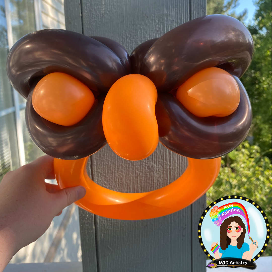 Balloon sculpture resembling a cartoon face with orange and brown balloons, including a large orange balloon for a nose, orange balloons for ears, and a brown balloon for hair, held by a person's hand against a background of trees and a building.