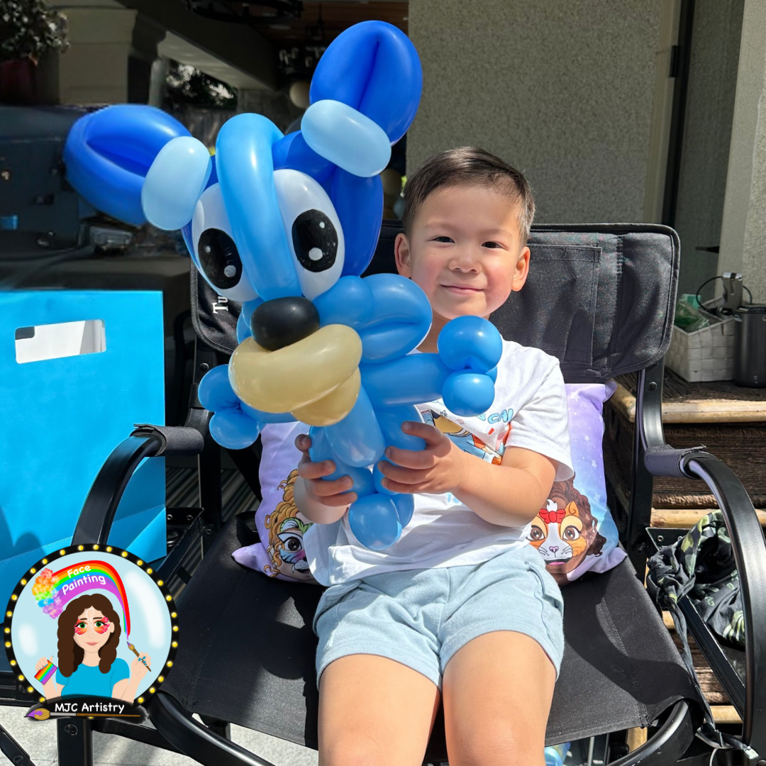 A young boy sitting in a chair outdoors, holding a balloon sculpture of Bluey made of blue, black, and beige balloons, smiling at the camera. Balloon twisting at a Vancouver birthday party.