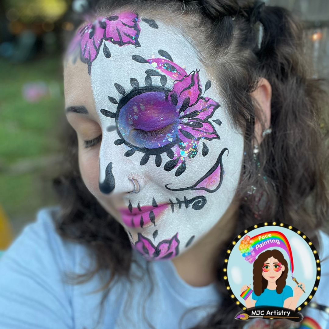 A woman with face paint resembling a Day of the Dead sugar skull, with colorful purple, pink, and black designs on her face, featuring floral and skull motifs, and glitter accents.
