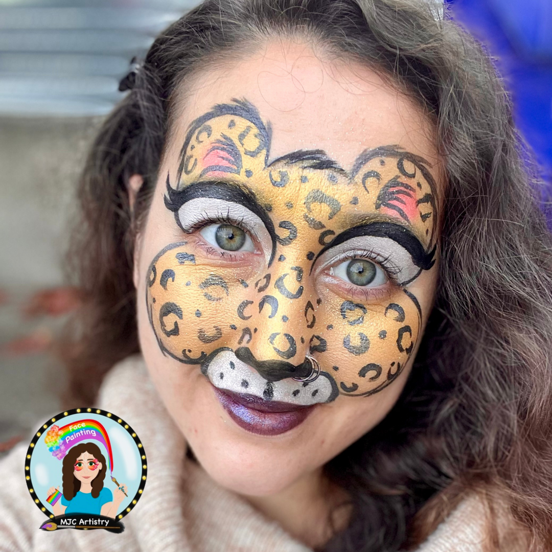 Woman with face painted as a leopard, featuring yellow and black spots, white accents around eyes, and a black nose with white spots. She has curly brown hair and light-colored eyes.