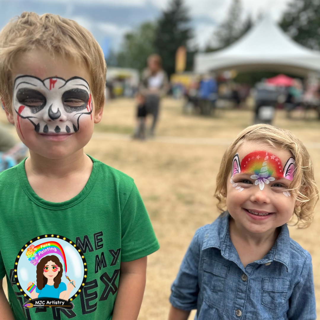 Two children with face paint, smiling at an outdoor event. The older boy on the left has a sugar skull face design, and the younger girl on the right has a rainbow unicorn face design. Tents and people are in the background.