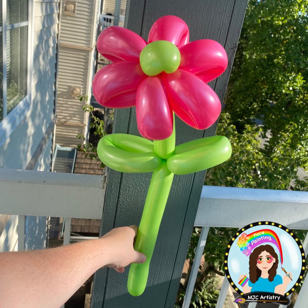 Colorful balloon sculpture depicting a flower with pink petals, a green center, and a green stem and leaves, held by a person's hand outdoors.