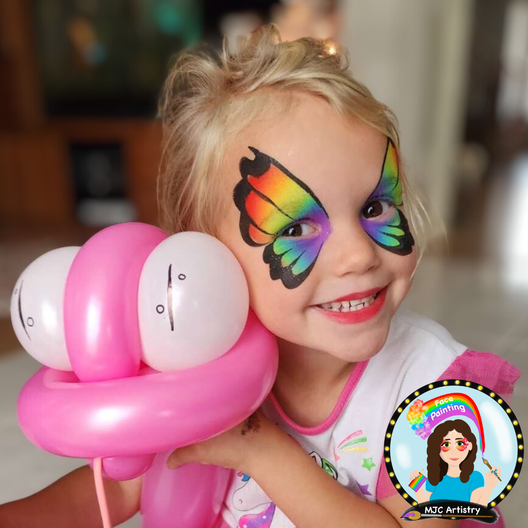 A young girl with rainbow butterfly face paint, smiling, holding pink balloons with smiley faces, take at a birthday party in Vancouver. 