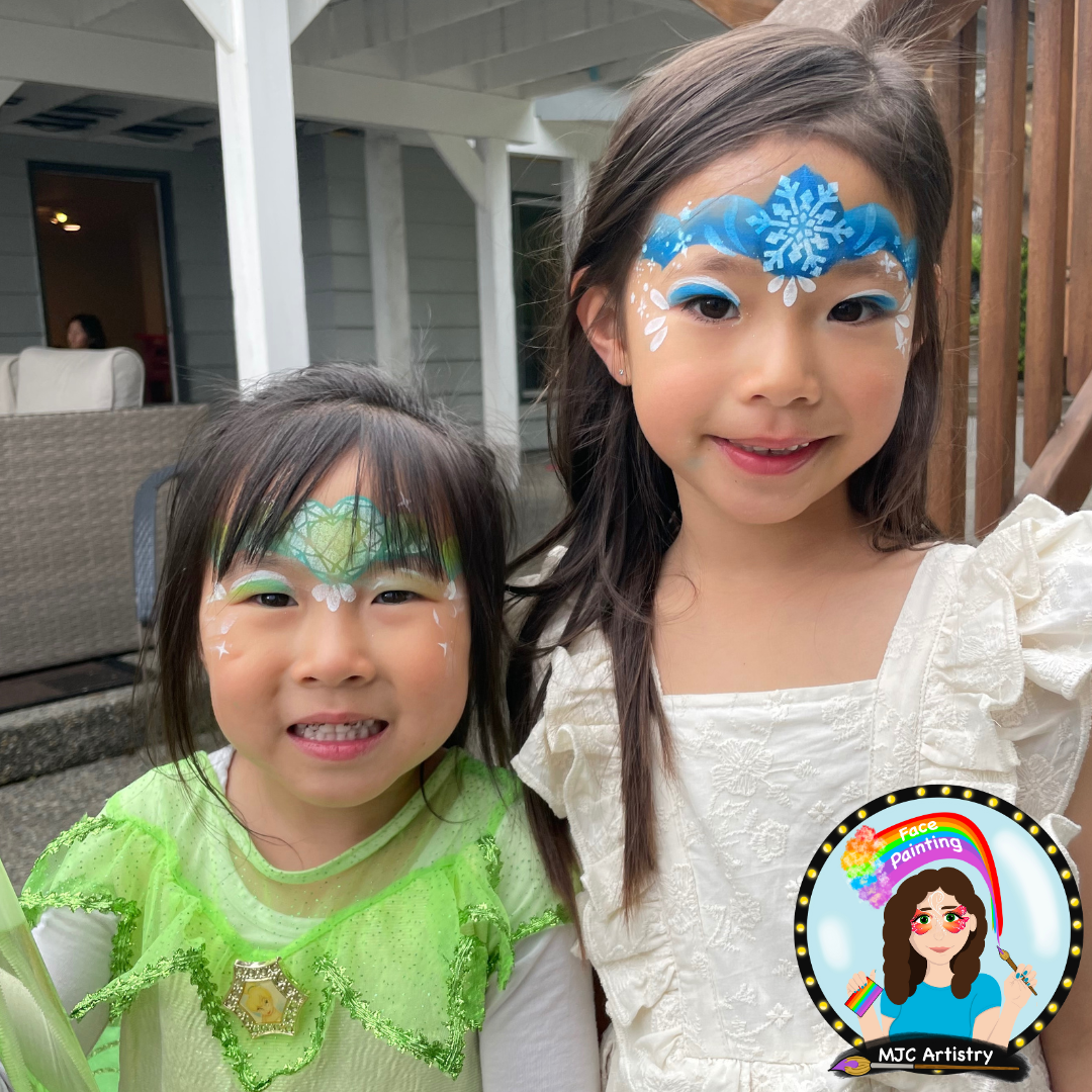 Two young girls with face paint, standing outside on a porch. The girl on the right has blue and white snowflake face paint, and the girl on the left has green and white face paint with a shamrock design. They are smiling at the camera.