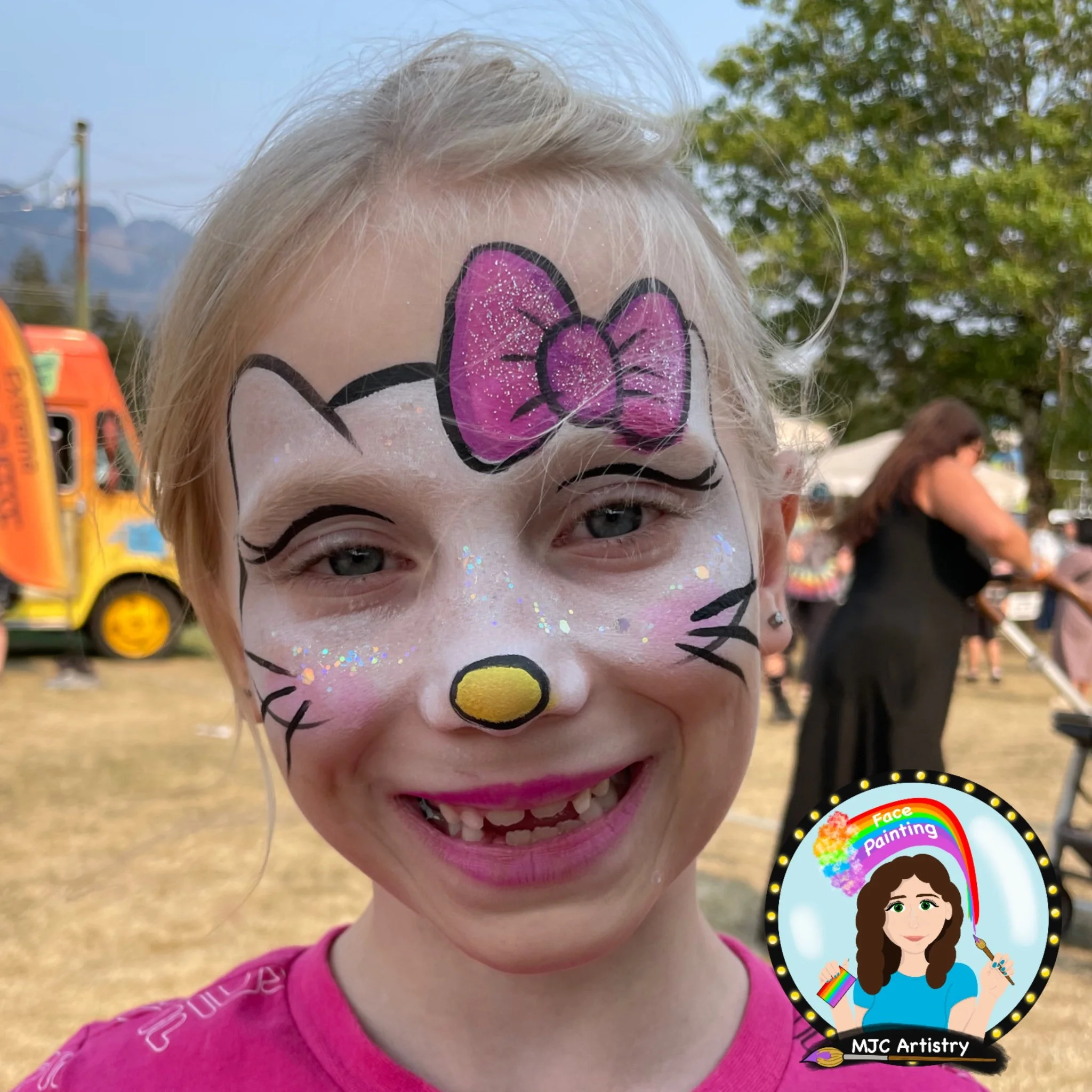 Young girl with face painted as Hello Kitty, featuring a pink bow, black whiskers, a yellow nose, and sparkles on her cheeks, smiling at an outdoor event.