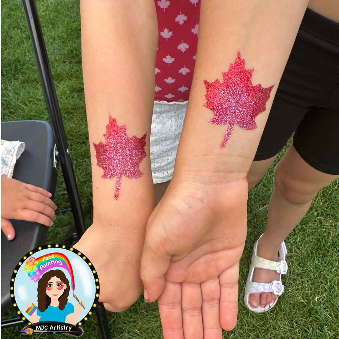 Two arms with matching pink glitter tattoos of maple leaves, held together outdoors on green grass.
