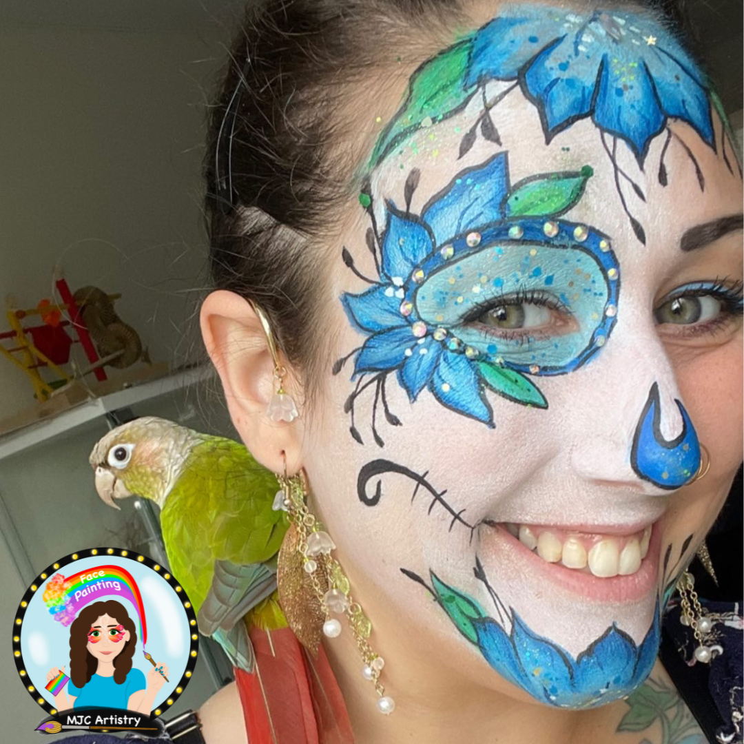 A smiling woman with painted face art and Halloween accessories. Her face painting features blue flowers, leaves, and decorative elements. She has earrings with pearl and leaf charms, and a green parakeet perches on her shoulder.
