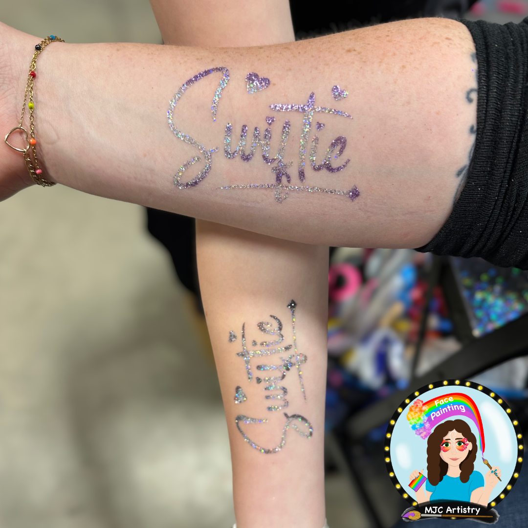 Two arms crossed, showcasing glittery temporary tattoos. One tattoo reads 'swiftie' in purple and pink glitter with hearts taken at a community event in Vancouver BC. 