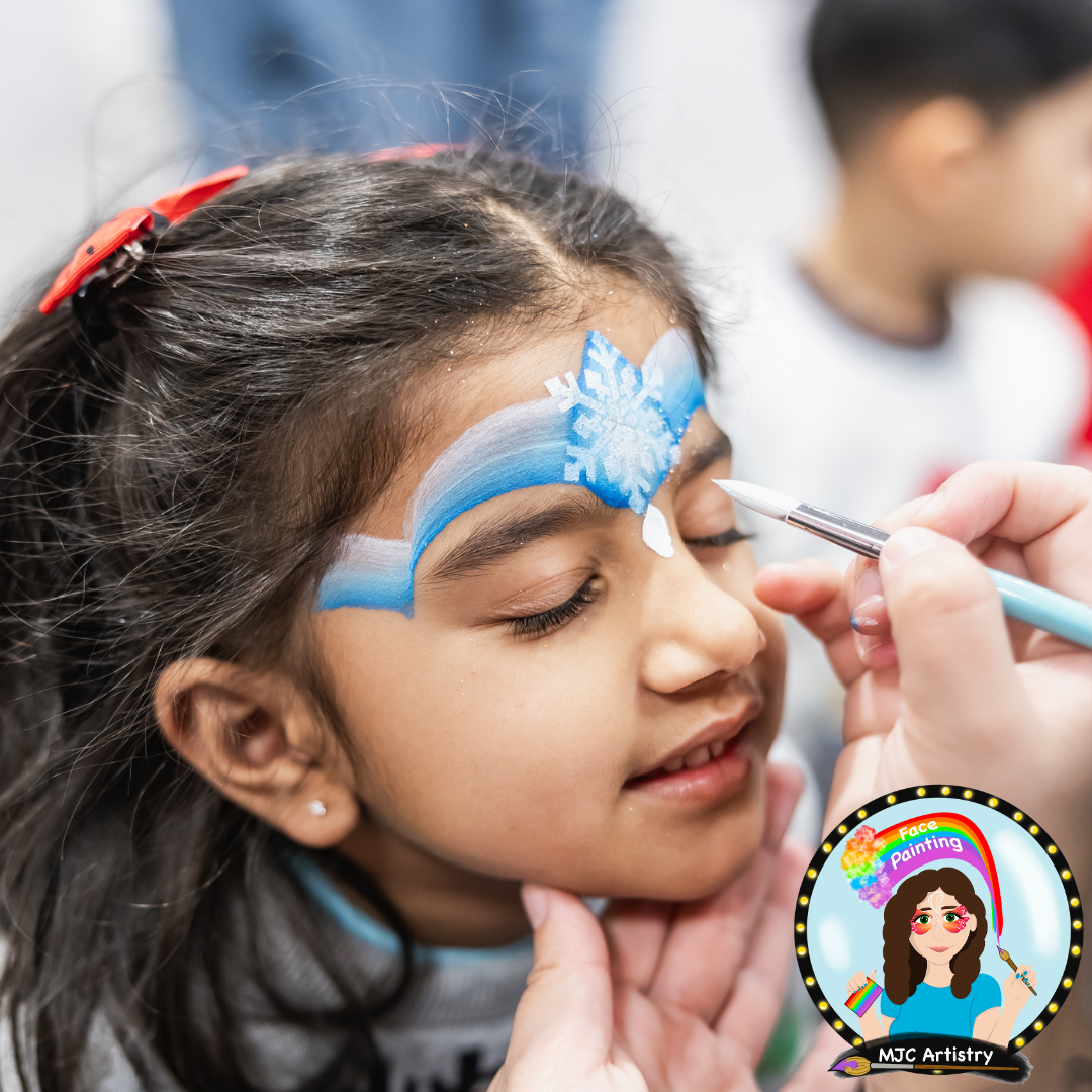A young girl with dark hair and a red hair clip is getting her face painted with a snowflake design in blue and white colours, with her eyes closed while the artist adds details at an event in Vancouver BC.